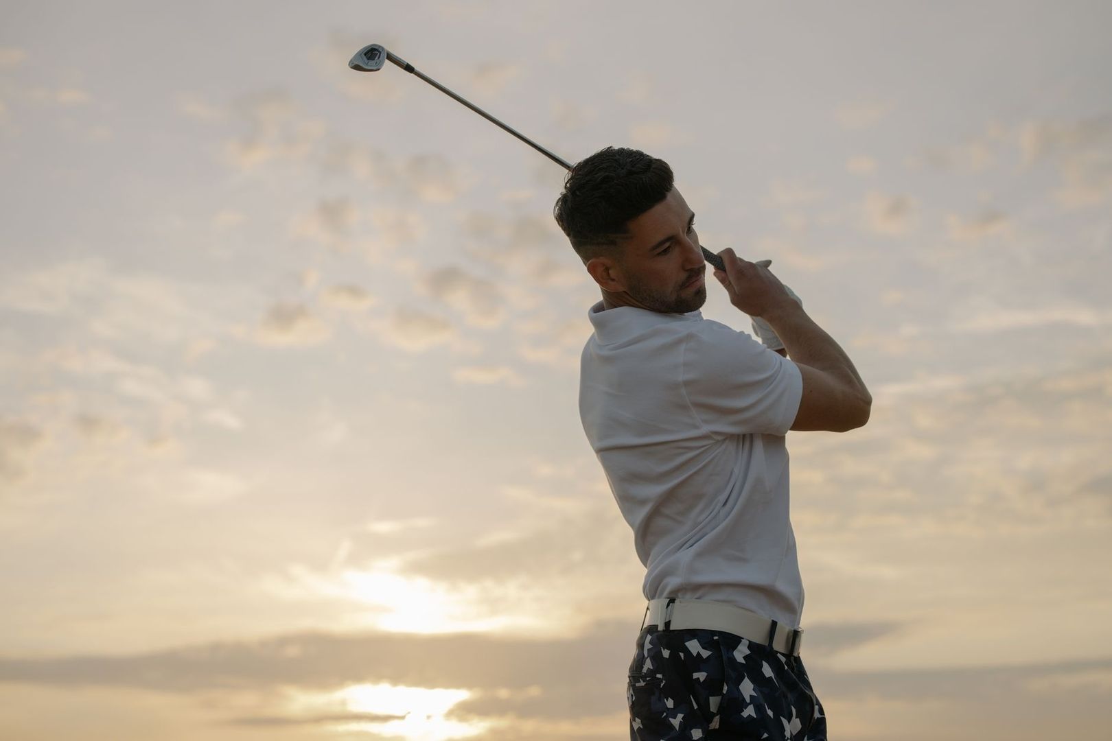 Golfer completing a swing against a cloudy, breezy-looking sunset sky